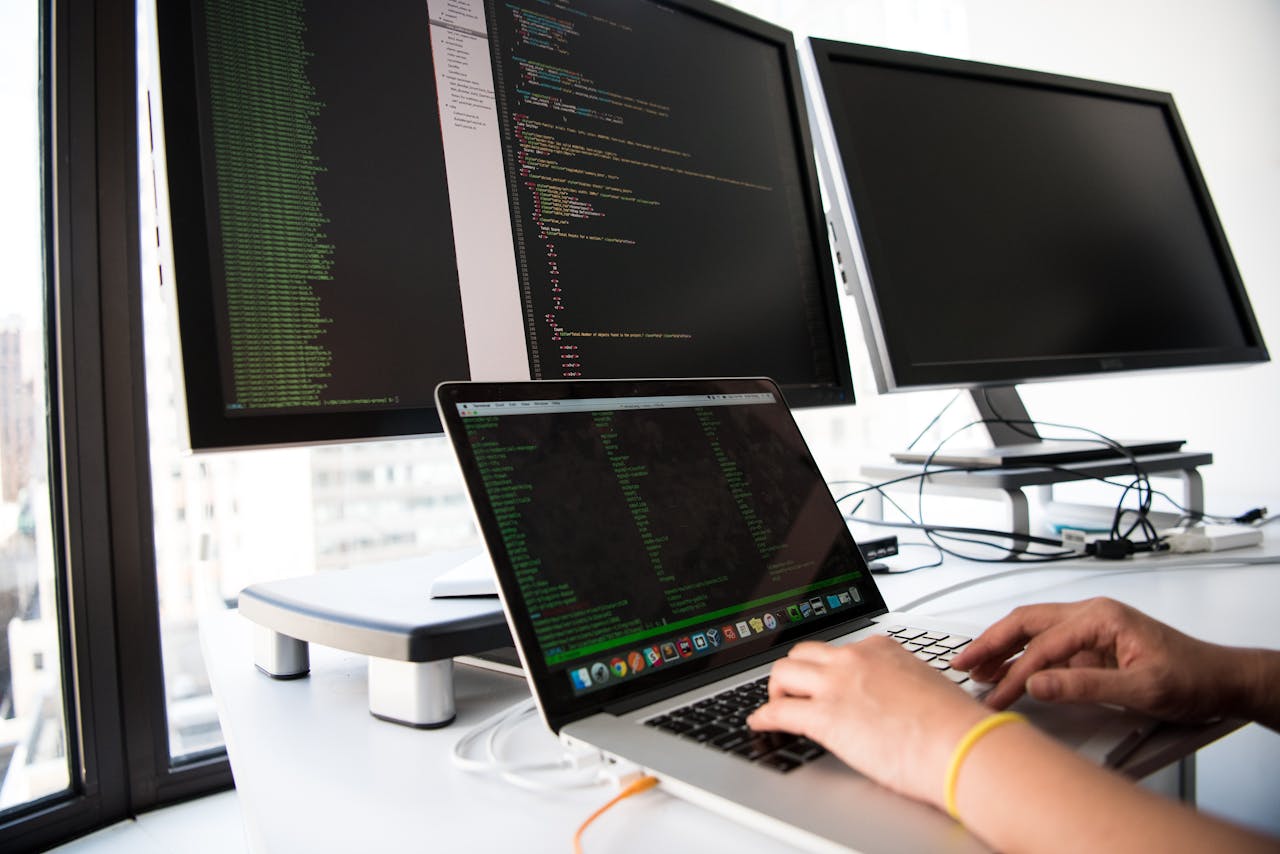 Home A developer writes code on a laptop in front of multiple monitors in an office setting.