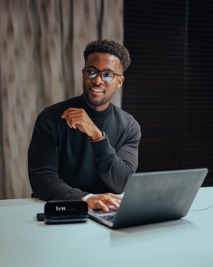 Home Cheerful man with glasses working on laptop at a table indoors, emanating a professional and modern workspace vibe.